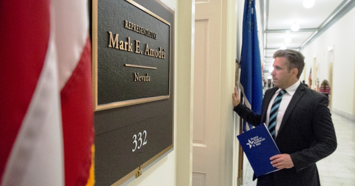 Young man in suit stands at a door in the distance. In front of shot, Mark Amodei's congressional door placard