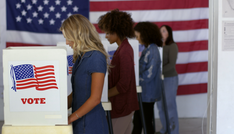 Photograph of people standing in front of voting booths in front of an American flag.