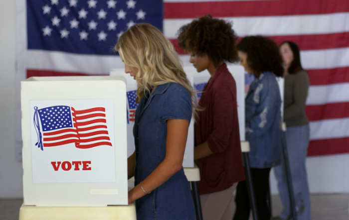 Four women cast ballots at voting booths with an American flag in the background.